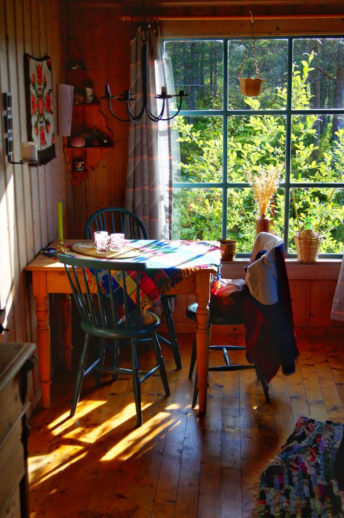 Sunlit rustic dining area with wooden furniture and vibrant decor overlooking greenery through a large window.